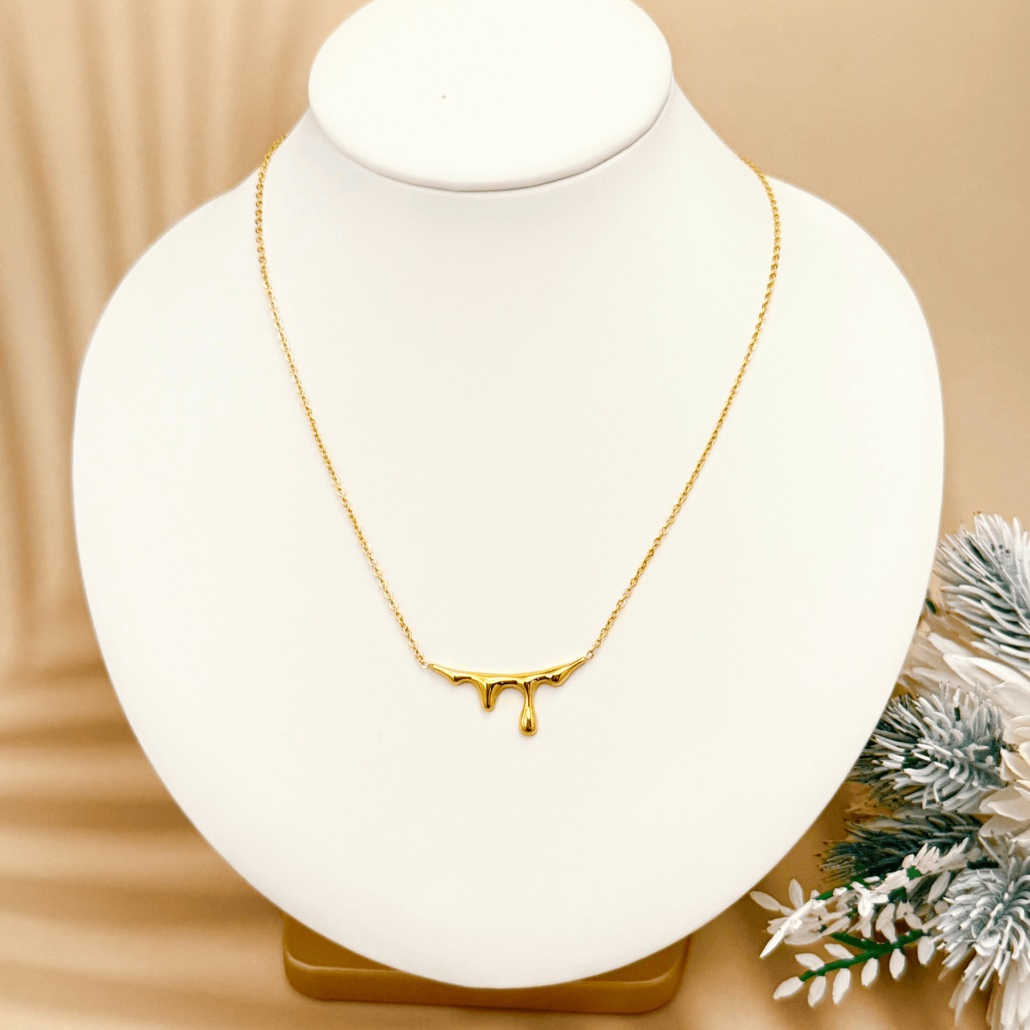 Gold drip-style pendant necklace with a sleek molten design on a fine chain, displayed on a white bust with soft floral accents.