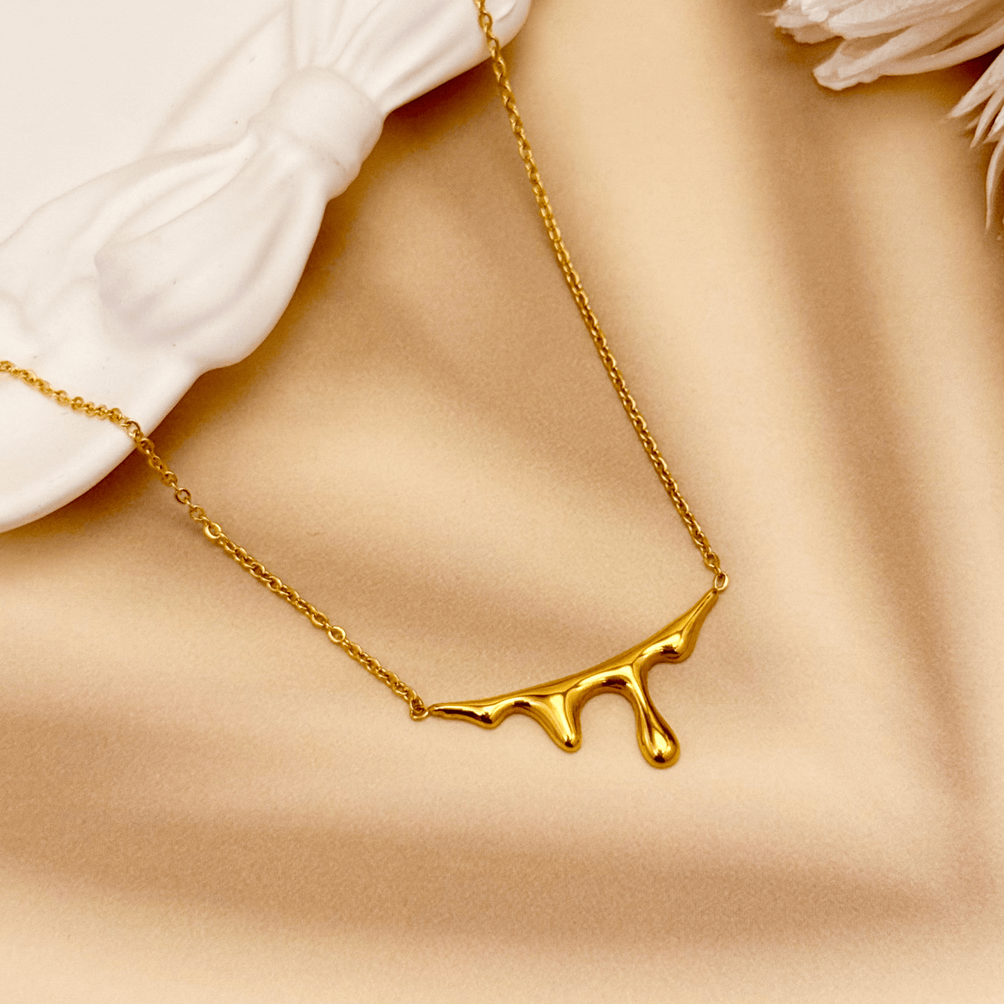Gold drip-style pendant necklace with a sleek molten design on a fine chain, displayed on a white bust with soft floral accents.