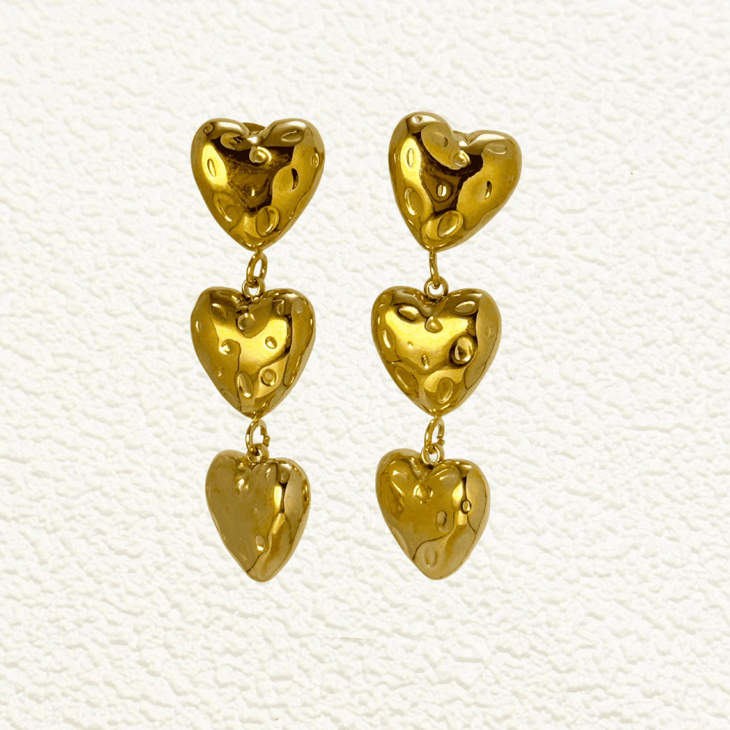Gold triple heart drop earrings made of textured PVD-plated stainless steel, displayed on a white textured background.