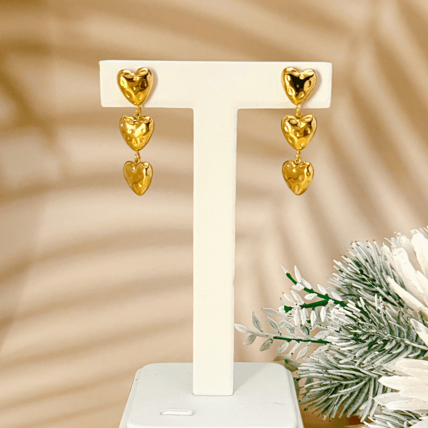 Gold triple heart drop earrings made of textured PVD-plated stainless steel, displayed on a white textured background.