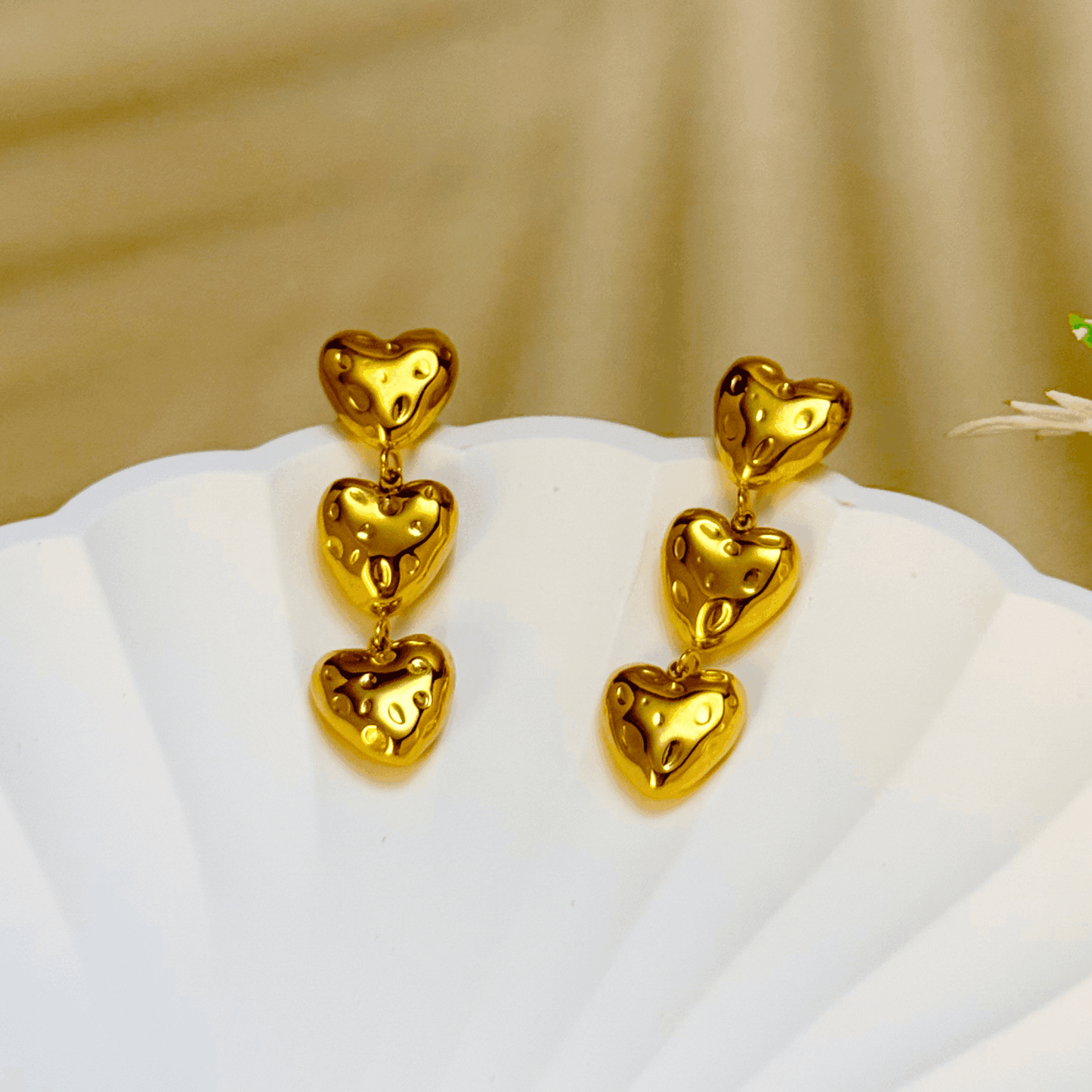Gold triple heart drop earrings made of textured PVD-plated stainless steel, displayed on a white textured background.