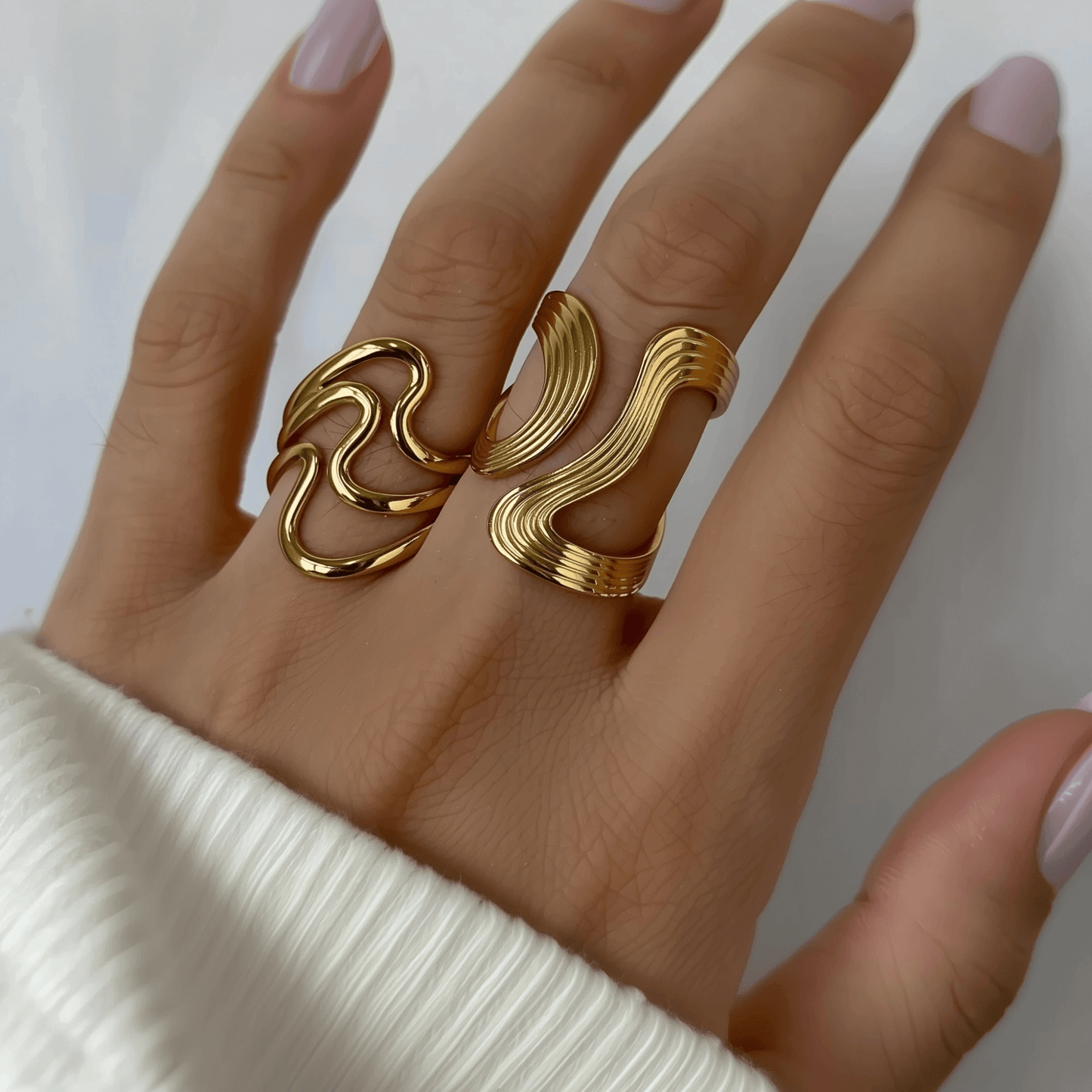 Gold sculptural ripple wave ring with layered curves, photographed on soft neutral background with floral decor.