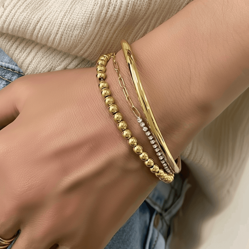 A close-up of a hand wearing a stacked gold bracelet collection, featuring a mix of polished bangle, beaded bracelet, dainty chain, and zircon-accented designs—perfect for stylish layering and everyday elegance.
