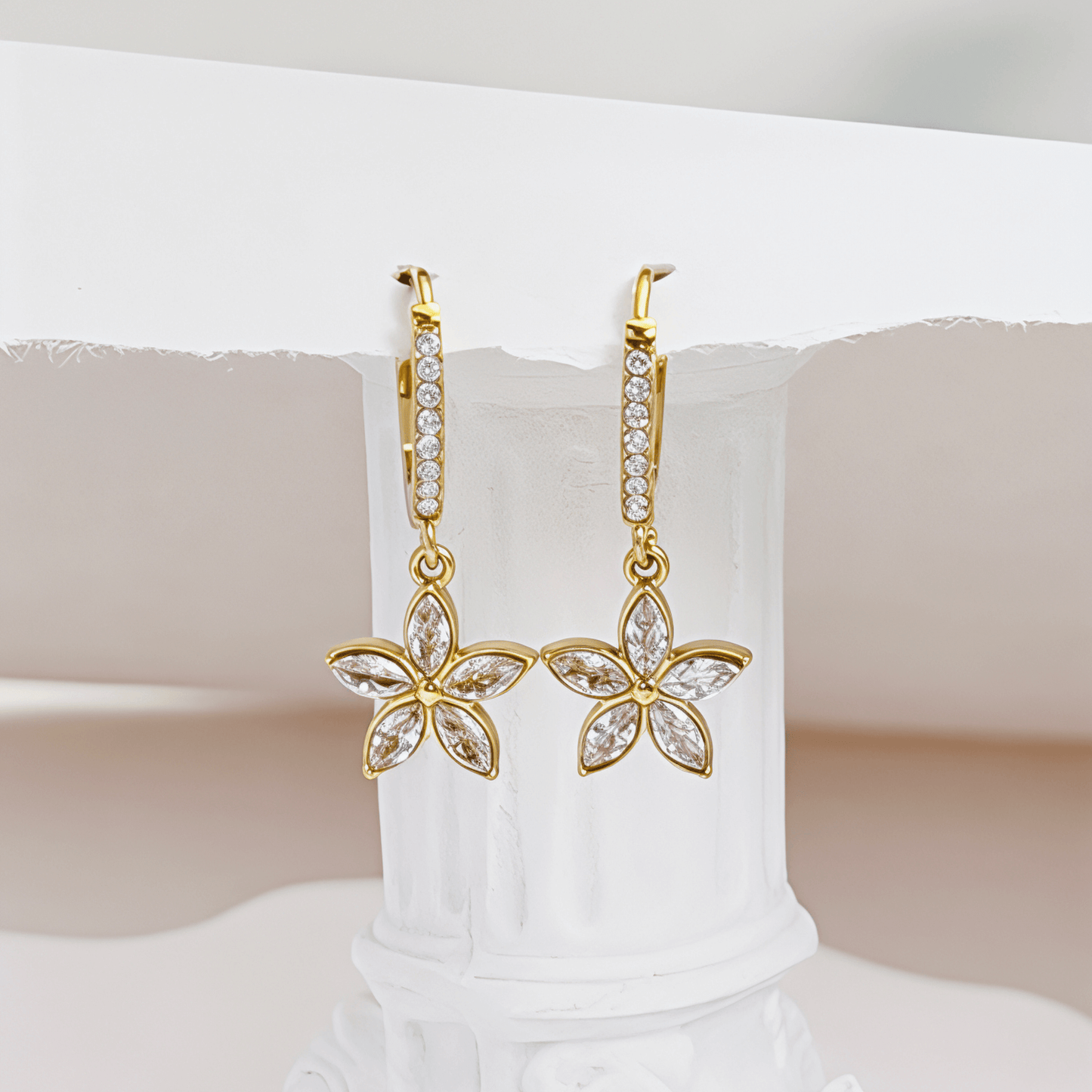 Gold huggie earrings featuring a crystal-lined hoop and a five-petalled flower charm by MXW Jewellery, displayed on a white textured surface.