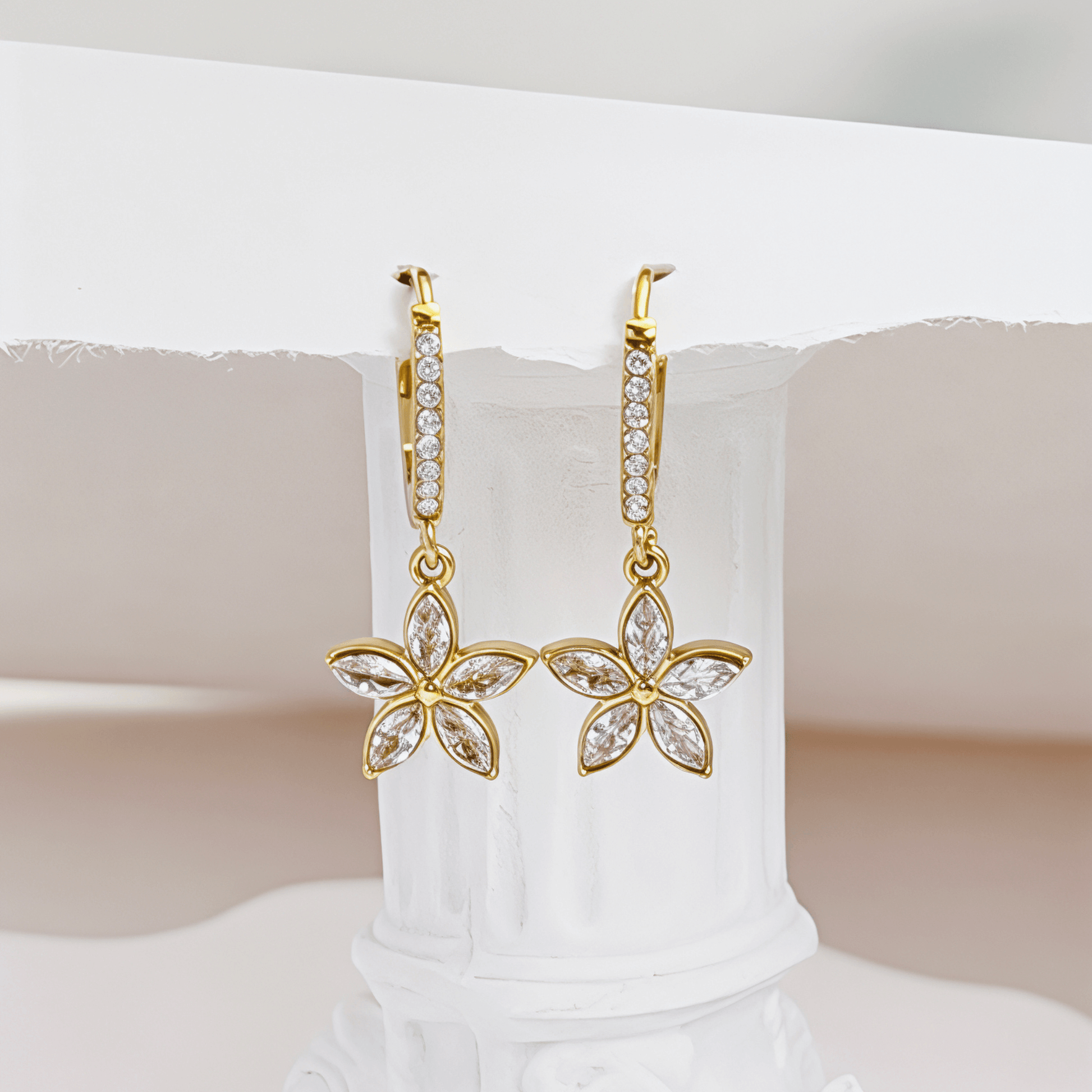 Gold huggie earrings featuring a crystal-lined hoop and a five-petalled flower charm by MXW Jewellery, displayed on a white textured surface.