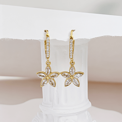 Gold huggie earrings featuring a crystal-lined hoop and a five-petalled flower charm by MXW Jewellery, displayed on a white textured surface.