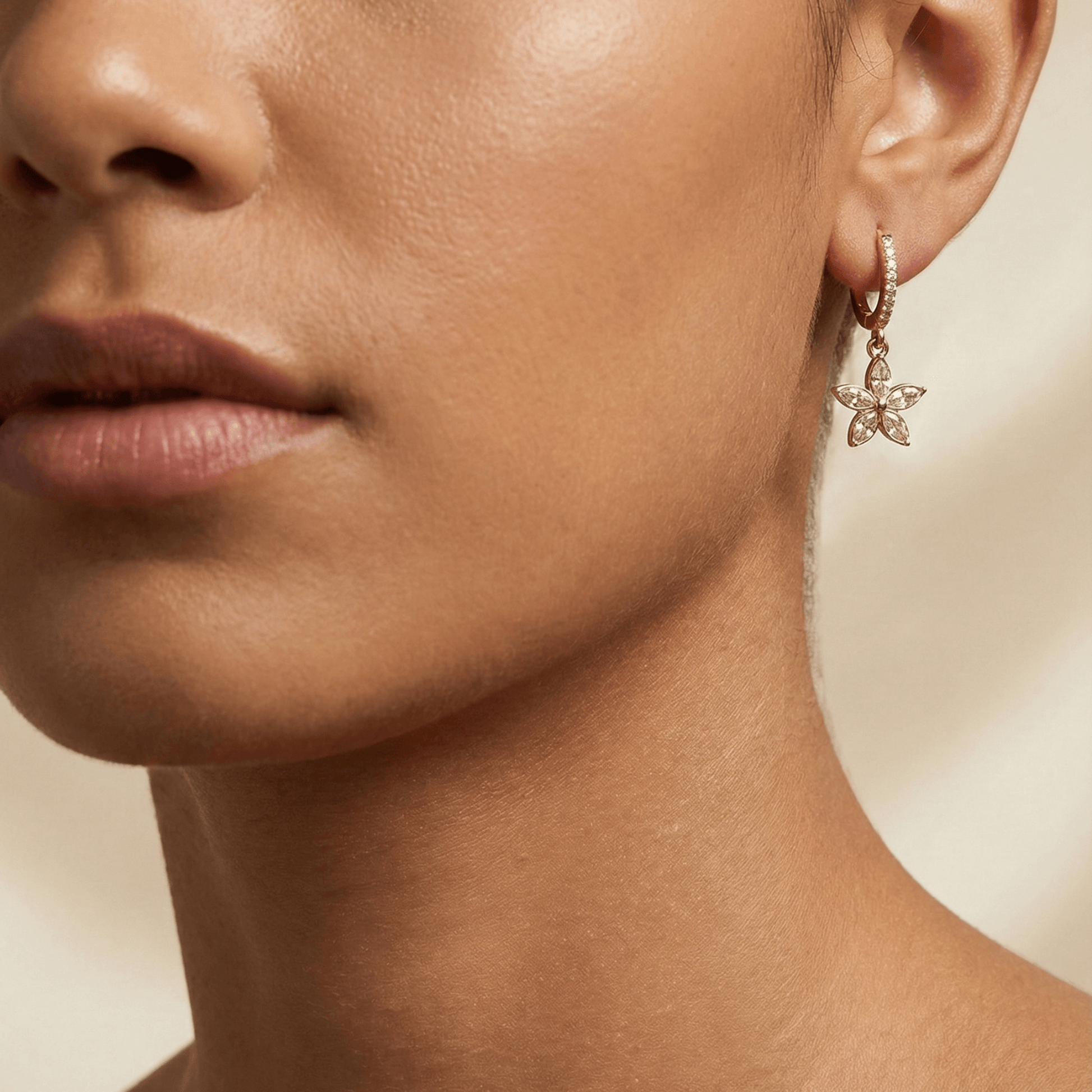 Gold huggie earrings featuring a crystal-lined hoop and a five-petalled flower charm by MXW Jewellery, displayed on a white textured surface.