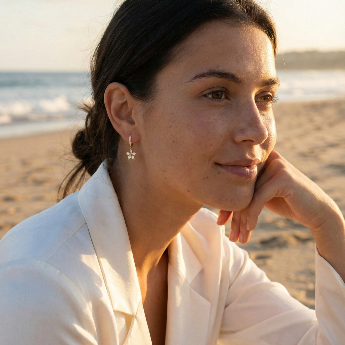 Gold huggie earrings featuring a crystal-lined hoop and a five-petalled flower charm by MXW Jewellery, displayed on a white textured surface.