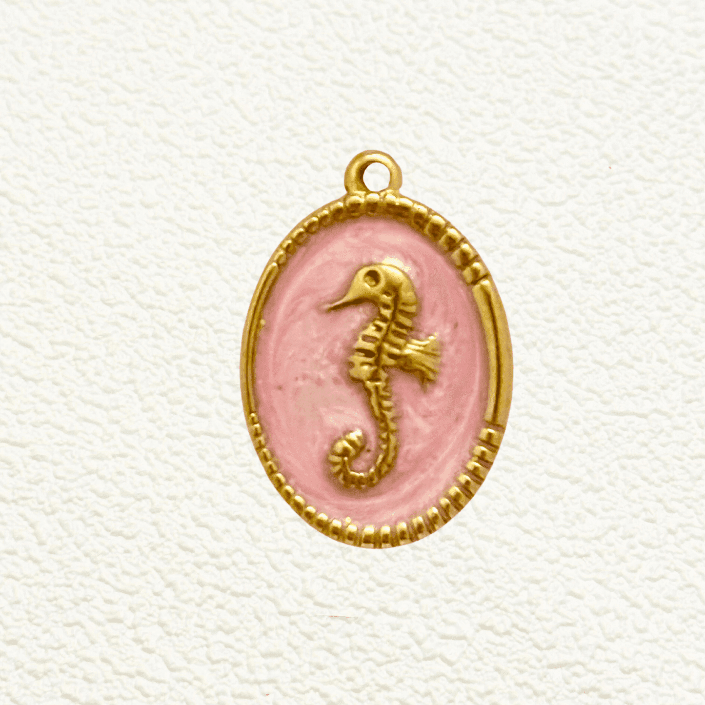 Gold seahorse charm set in an oval medallion with a soft blush pink enamel background and detailed golden beaded edge.