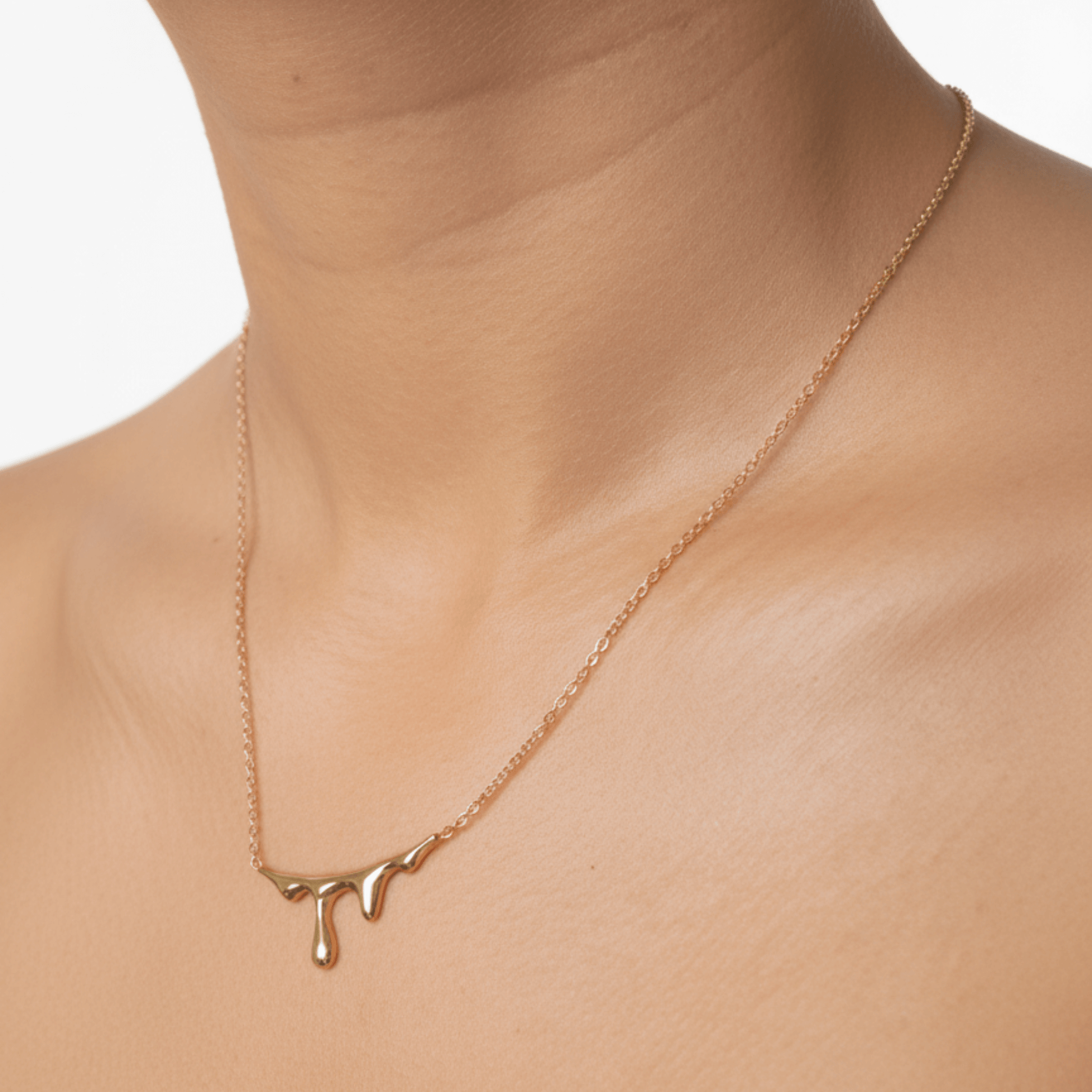 Gold drip-style pendant necklace with a sleek molten design on a fine chain, displayed on a white bust with soft floral accents.