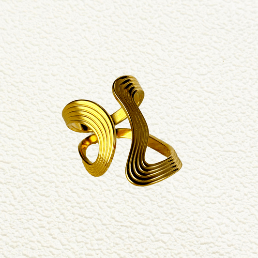 Gold sculptural ripple wave ring with layered curves, photographed on soft neutral background with floral decor.