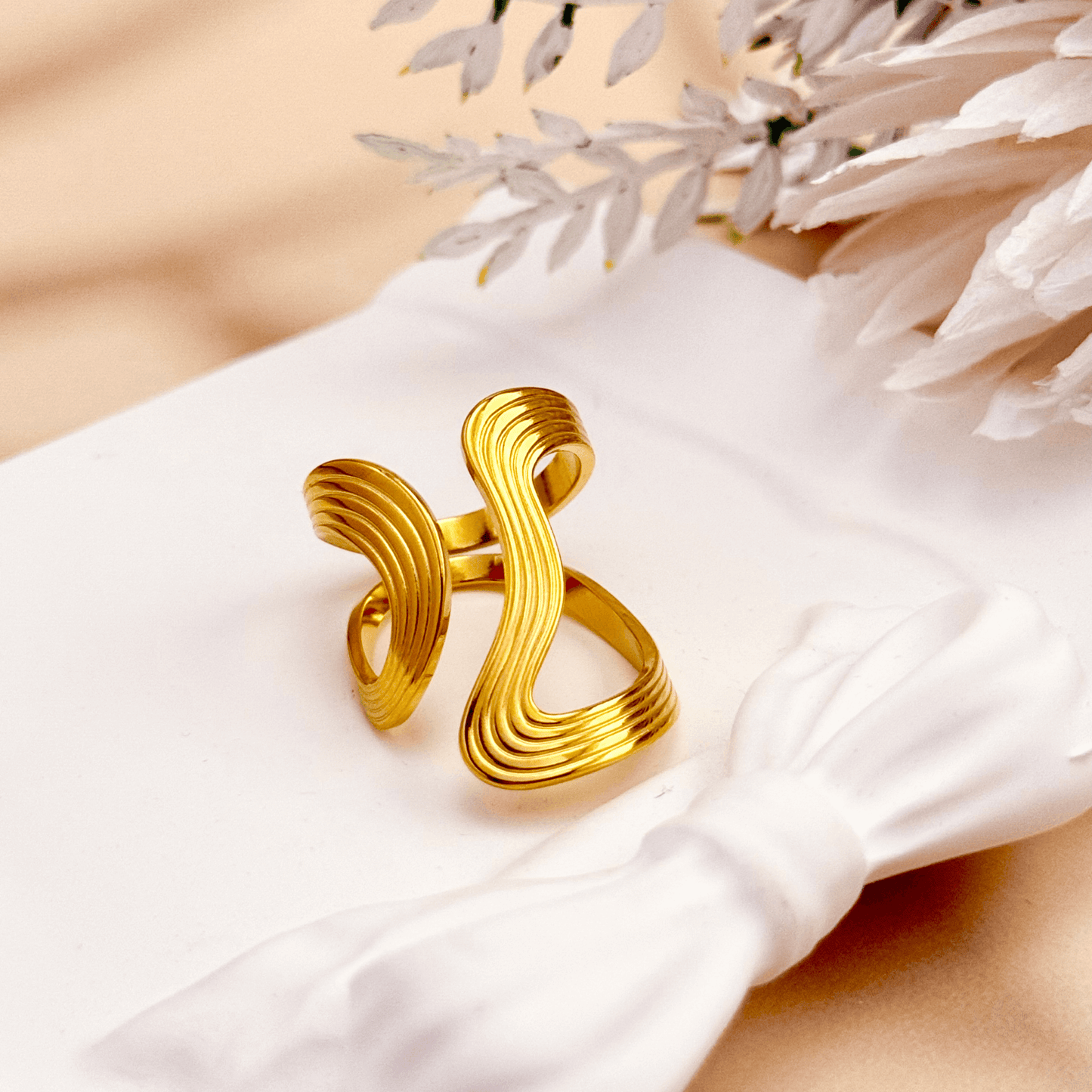 Gold sculptural ripple wave ring with layered curves, photographed on soft neutral background with floral decor.