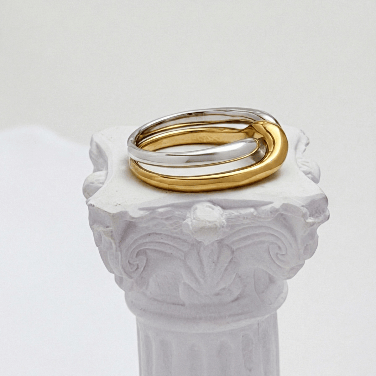 Two-Tone Interlocking Ring by MXW Jewellery featuring interwoven gold and silver bands displayed on a white textured pedestal.