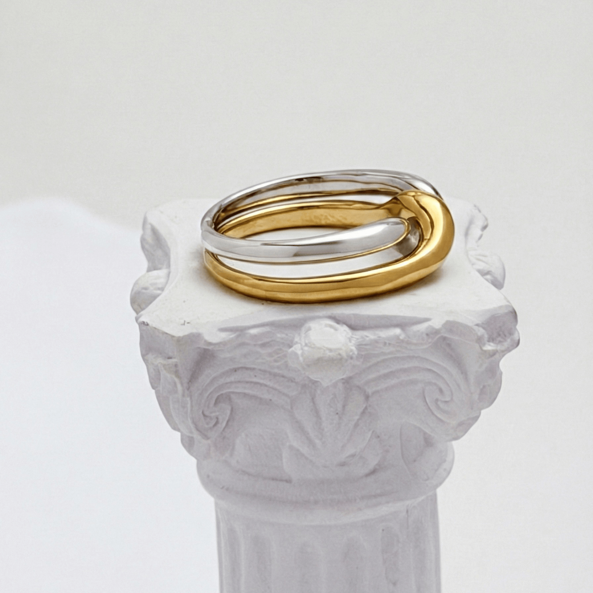 Two-Tone Interlocking Ring by MXW Jewellery featuring interwoven gold and silver bands displayed on a white textured pedestal.