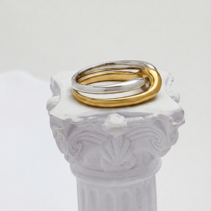 Two-Tone Interlocking Ring by MXW Jewellery featuring interwoven gold and silver bands displayed on a white textured pedestal.