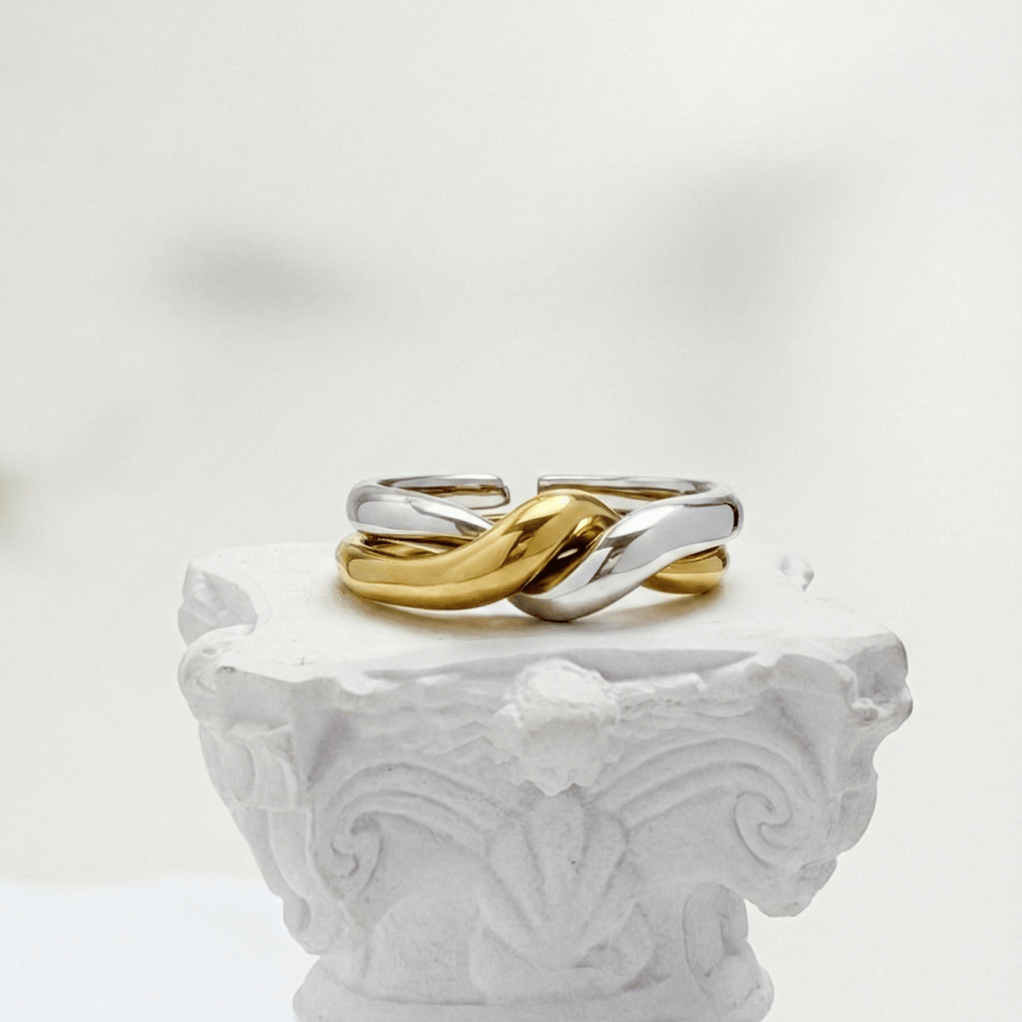 Two-Tone Interlocking Ring by MXW Jewellery featuring interwoven gold and silver bands displayed on a white textured pedestal.