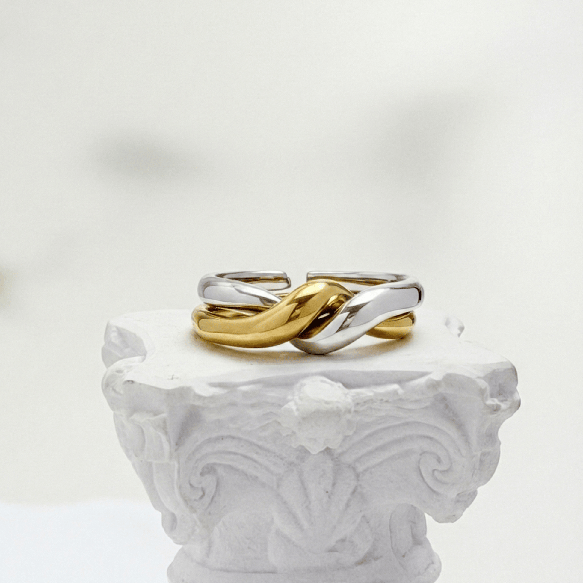 Two-Tone Interlocking Ring by MXW Jewellery featuring interwoven gold and silver bands displayed on a white textured pedestal.